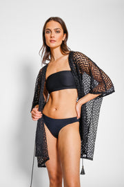 Brunette model facing front close up, wearing a Black Zuma Crochet Beach Kimono. This kimono features a sheer crochet with a zig-zag herringbone pattern and a tassel tie, perfect for lounging, pool-side hangouts, and summer strolls. The loose fit ensures a light and breezy feel. Made of 100% polyester. Koy Resort affordable vacation, cruise, and resort-wear.