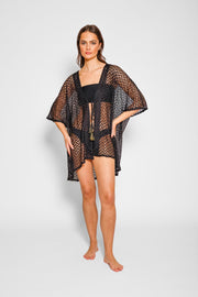 Brunette model facing front, wearing a Black Zuma Crochet Beach Kimono. This kimono features a sheer crochet with a zig-zag herringbone pattern and a tassel tie, perfect for lounging, pool-side hangouts, and summer strolls. The loose fit ensures a light and breezy feel. Made of 100% polyester. Koy Resort affordable vacation, cruise, and resort-wear.