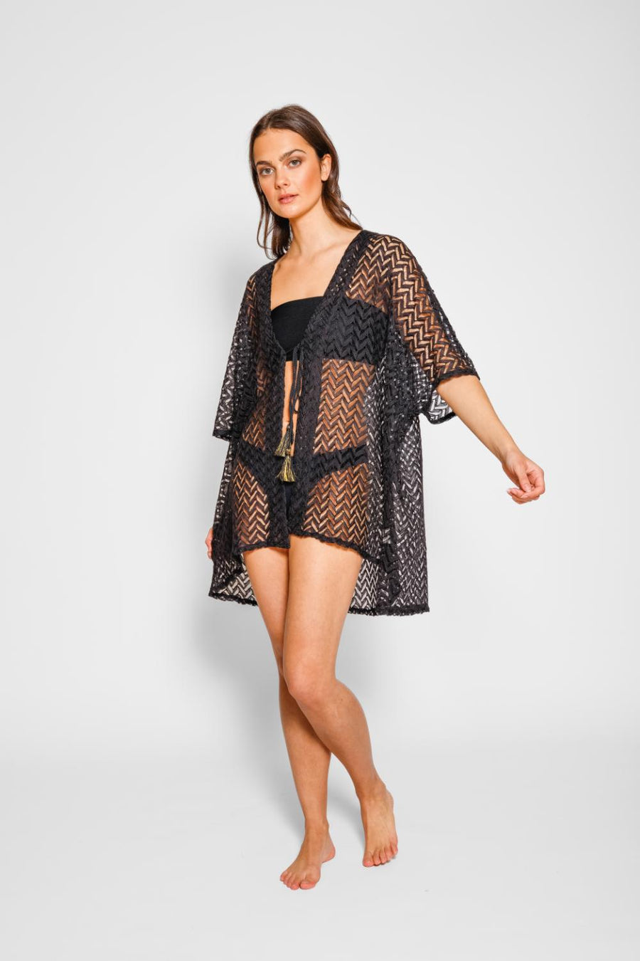 Brunette model facing front, wearing a Black Zuma Crochet Beach Kimono. This kimono features a sheer crochet with a zig-zag herringbone pattern and a tassel tie, perfect for lounging, pool-side hangouts, and summer strolls. The loose fit ensures a light and breezy feel. Made of 100% polyester. Koy Resort affordable vacation, cruise, and resort-wear.