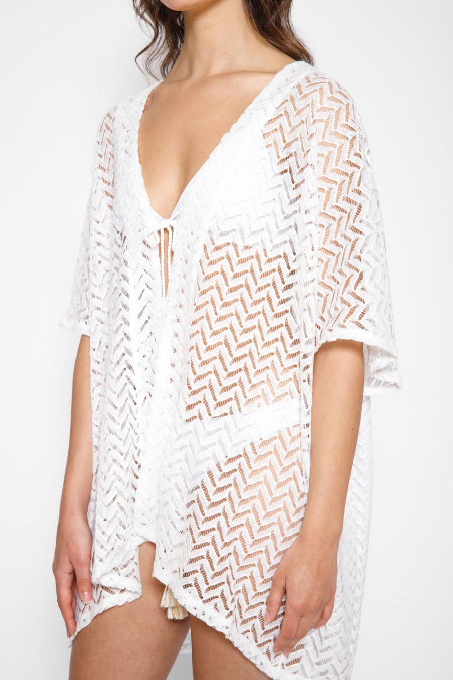 Brunette model facing front close up, wearing a Natural White Zuma Crochet Beach Kimono. This kimono features a sheer crochet with a zig-zag herringbone pattern and a tassel tie, perfect for lounging, pool-side hangouts, and summer strolls. The loose fit ensures a light and breezy feel. Made of 100% polyester. Koy Resort affordable vacation, cruise, and resort-wear.