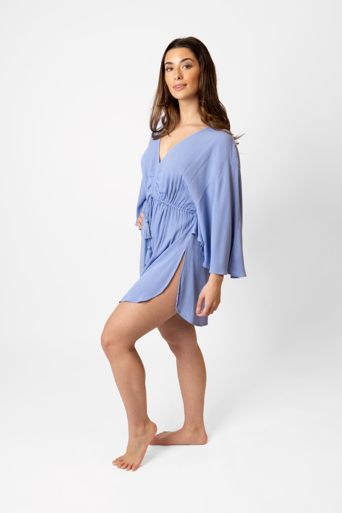 Koy resort miami cinched front beachy batwing mini dress. Brunette model facing side, wearing Bahama Blue Miami Cinched Front Dress. Features ruched center front with tassel pull and gold trim, adjustable straps, side slits, and butterfly batwing style sleeves. Light and breezy fabric, perfect for making a statement in South Beach. Koy Resort affordable vacation, cruise, and resort-wear.