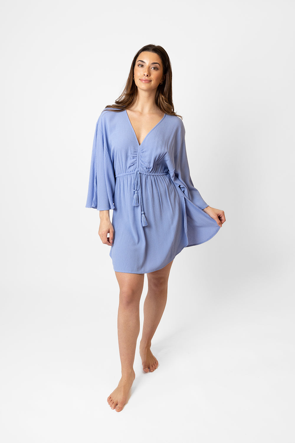 Koy resort miami cinched front beachy batwing mini dress. Brunette model facing front, wearing Bahama Blue Miami Cinched Front Dress. Features ruched center front with tassel pull and gold trim, adjustable straps, side slits, and butterfly batwing style sleeves. Light and breezy fabric, perfect for making a statement in South Beach. Koy Resort affordable vacation, cruise, and resort-wear.