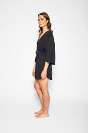 Brunette model facing side, wearing Black Miami Cinched Front Dress with butterfly sleeves, ruched center front with a tassel pull detail, adjustable straps, and side slits. Koy Resort affordable vacation, cruise, and resort-wear.