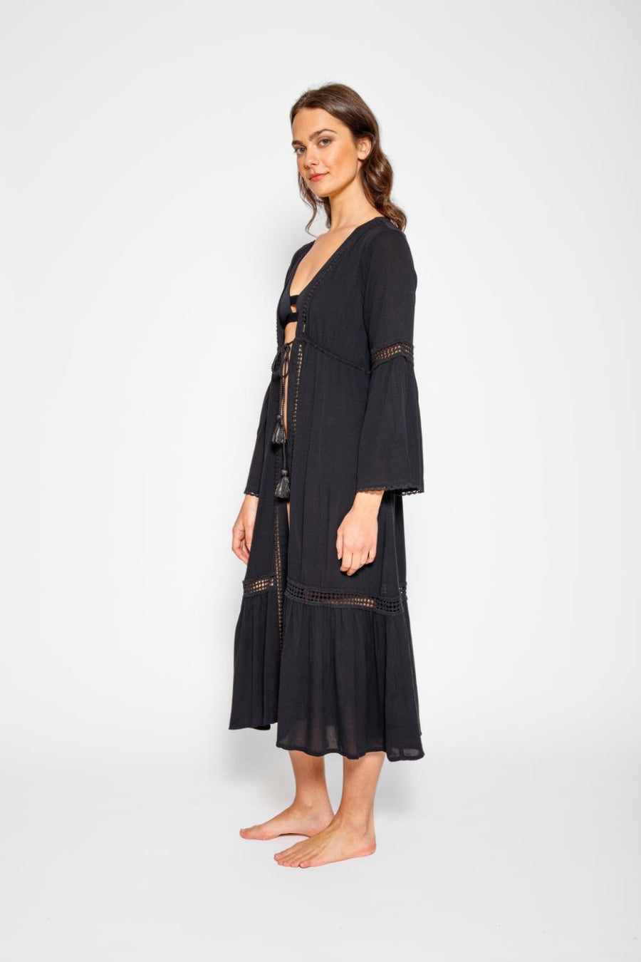 Brunette model facing side, wearing Black Miami Luxe Long Sleeve Robe with bell sleeves, maxi length, and tie front. Features crochet detailing, luxury lace trim, and pockets. Made of 100% crinkle rayon. Perfect for a sophisticated and comfortable look. Koy Resort affordable vacation, cruise, and resort-wear.