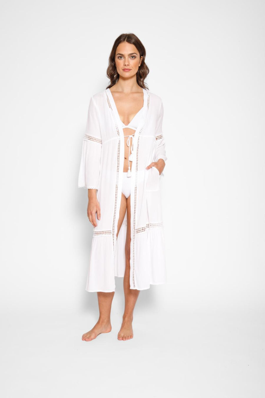 Brunette model facing front, wearing White Miami Luxe Long Sleeve Robe with bell sleeves, maxi length, and tie front. Features crochet detailing, luxury lace trim, and pockets. Made of 100% crinkle rayon. Perfect for a sophisticated and comfortable look. Koy Resort affordable vacation, cruise, and resort-wear.