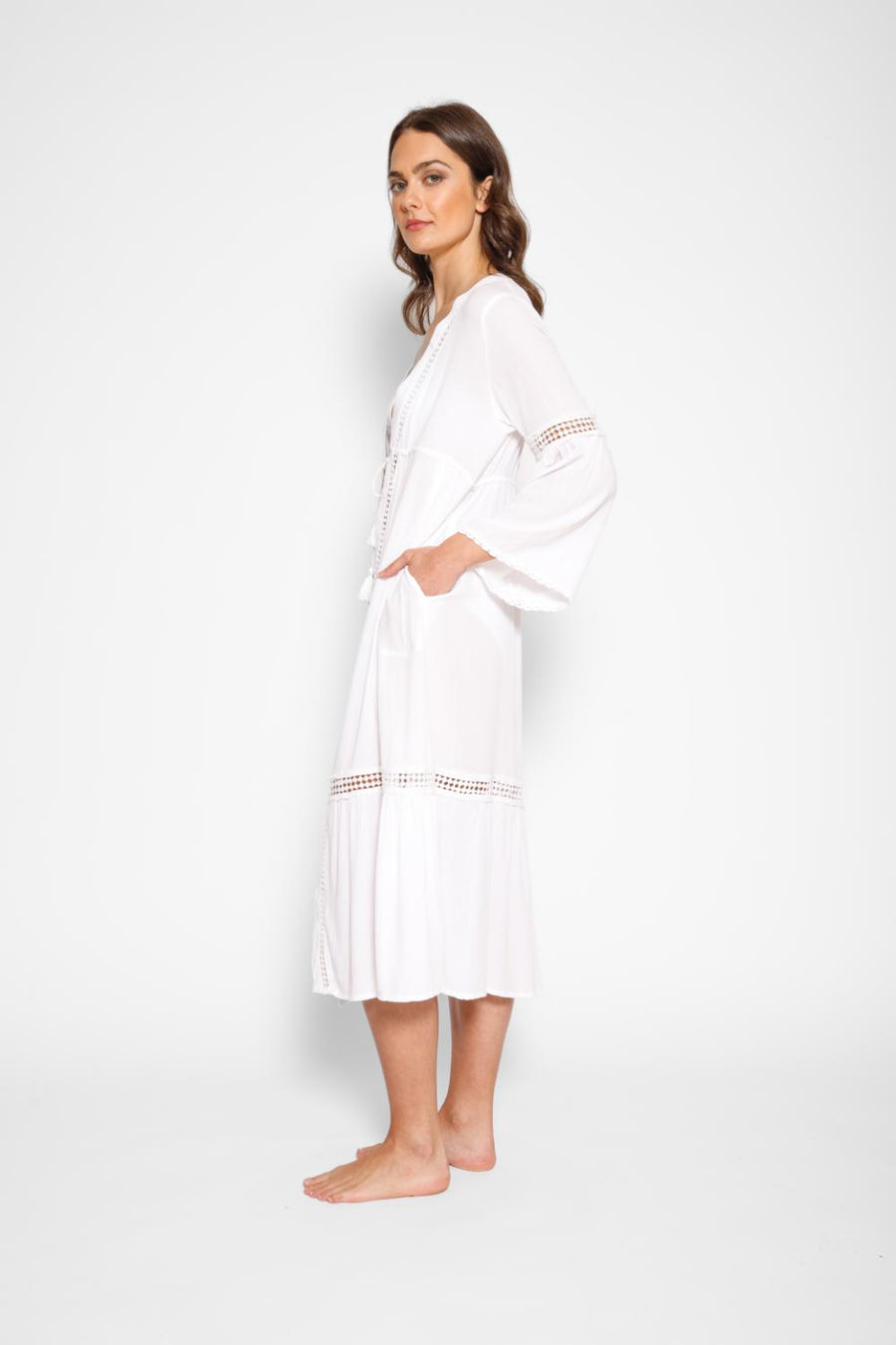 Brunette model facing side, wearing White Miami Luxe Long Sleeve Robe with bell sleeves, maxi length, and tie front. Features crochet detailing, luxury lace trim, and pockets. Made of 100% crinkle rayon. Perfect for a sophisticated and comfortable look. Koy Resort affordable vacation, cruise, and resort-wear.