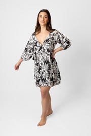 Brunette model facing side, wearing Black Onyx Copacabana Floral V-Neck Tunic Dress. Features a tropical hibiscus floral print, v-neck with tassel, and trapeze style. Made from 100% sand-washed rayon for a cool and comfortable feel, perfect for vacations or everyday wear. Koy Resort affordable vacation, cruise, and resort-wear.