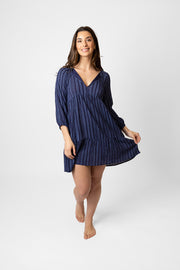 Brunette model facing front, wearing a navy blue Marina Stripe V-Neck Tunic Dress. Features a v-neck with tassel, mock stripe print, and elastic-trimmed long sleeves. Made from 100% crinkle rayon. Perfect for everyday wear with a touch of flair. Koy Resort affordable vacation, cruise, and resort-wear.