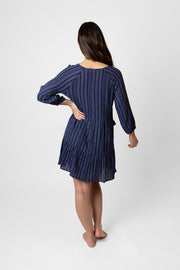 Brunette model facing back, wearing a navy blue Marina Stripe V-Neck Tunic Dress. Features a v-neck with tassel, mock stripe print, and elastic-trimmed long sleeves. Made from 100% crinkle rayon. Perfect for everyday wear with a touch of flair. Koy Resort affordable vacation, cruise, and resort-wear.