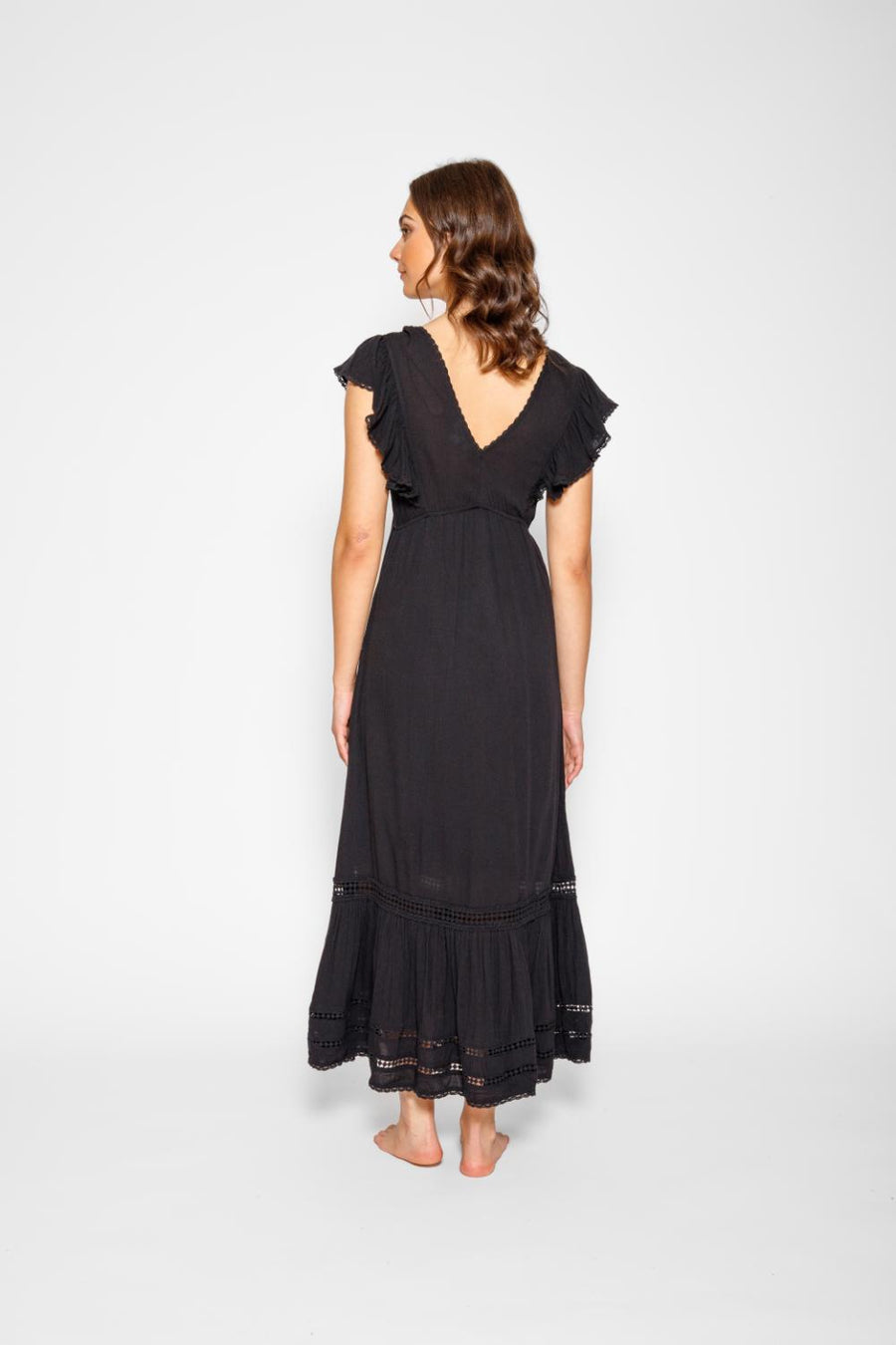 Brunette model facing back, wearing Black Miami Luxe Ruffle Sleeve Maxi Dress with ruffle sleeves, deep v-neck, semi-exposed back, empire waist, luxury crochet details, and frill hem. Koy Resort affordable vacation, cruise, and resort-wear.