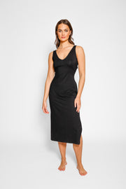 Brunette model facing front, wearing Black Laguna Beach Twist Dress with a twisted back with cutouts, scoop v-neck, asymmetrical midi length, and elegant hem. Made from soft cotton jersey for a figure-flattering and sophisticated look. Koy Resort affordable vacation, cruise, and resort-wear.