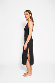 Brunette model facing side, wearing Black Laguna Beach Twist Dress with a twisted back with cutouts, scoop v-neck, asymmetrical midi length, and elegant hem. Made from soft cotton jersey for a figure-flattering and sophisticated look. Koy Resort affordable vacation, cruise, and resort-wear.