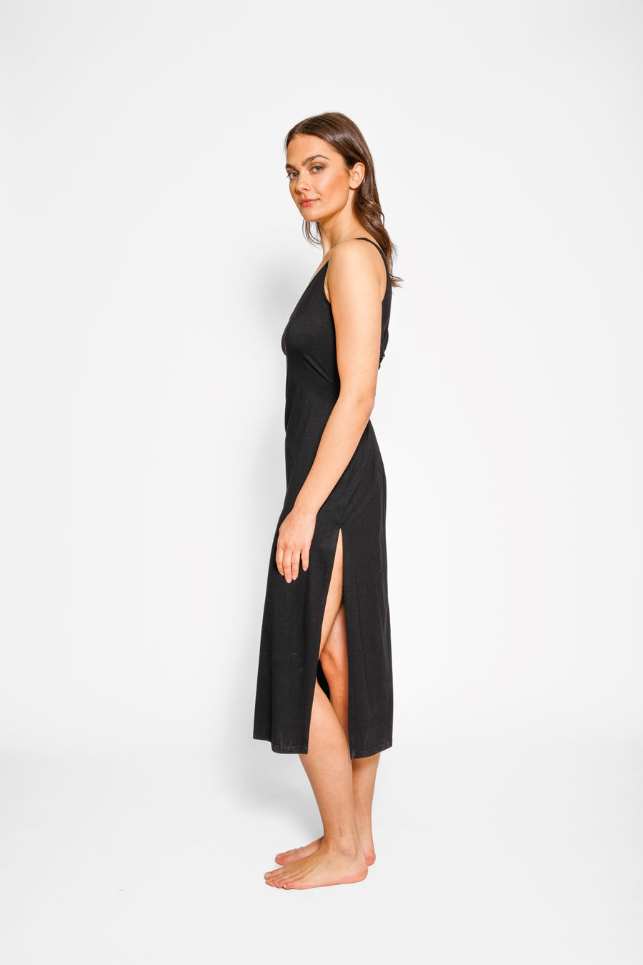 Brunette model facing side, wearing Black Laguna Beach Twist Dress with a twisted back with cutouts, scoop v-neck, asymmetrical midi length, and elegant hem. Made from soft cotton jersey for a figure-flattering and sophisticated look. Koy Resort affordable vacation, cruise, and resort-wear.