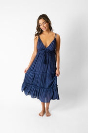 Brunette model facing front, wearing a navy Marina Stripe Strappy Maxi Dress. Features a deep V-neck, tiered design, and adjustable tassel trim chest. The dress is full length with an empire waist and mock stripe print. Perfect for staying comfy and stylish. Koy Resort affordable vacation, cruise, and resort-wear.