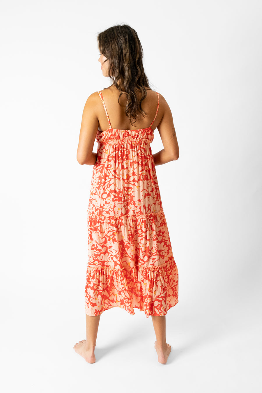 Brunette model facing back, wearing Pink Valencia Midi Dress with a pink floral print, deep v-neck, adjustable spaghetti straps, empire waist with tassel, tiered frill hem, and midi length. Koy Resort affordable vacation, cruise, and resort-wear.