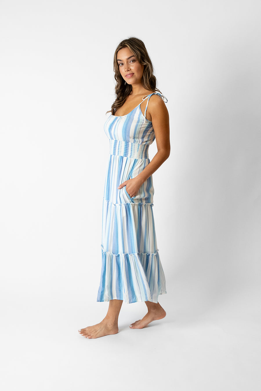 Brunette model facing side, wearing Taormina Blue Sicily Midi Dress with adjustable tie straps, scoop neck, empire wide elastic waist, tiers, and pockets. Koy Resort affordable vacation, cruise, and resort-wear.