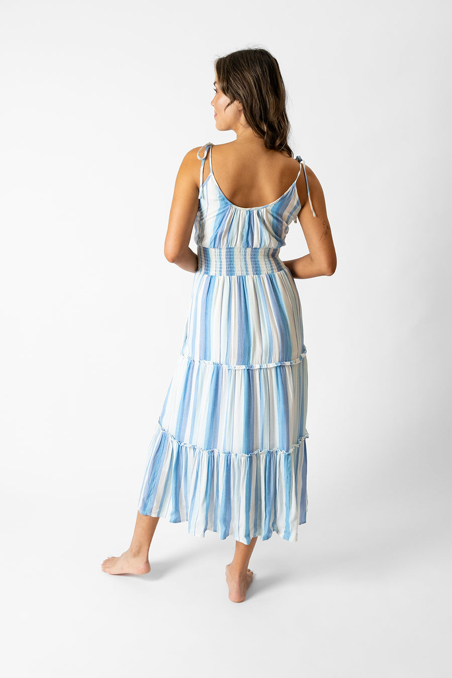 Brunette model facing back, wearing Taormina Blue Sicily Midi Dress with adjustable tie straps, scoop neck, empire wide elastic waist, tiers, and pockets. Koy Resort affordable vacation, cruise, and resort-wear.