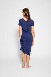 Brunette model facing back, wearing Navy Laguna Beach Midi Dress with adjustable side gathering, scoop neck, short sleeves, asymmetrical hem, and a figure-flattering fit. Made from a cozy cotton jersey blend for stylish and comfortable wear. Koy Resort affordable vacation, cruise, and resort-wear.