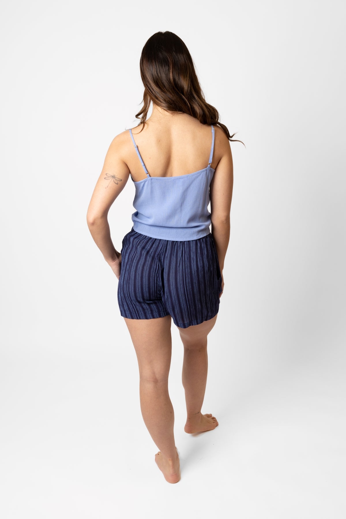 Brunette model facing back, wearing navy Marina Stripe Front Tie Shorts. Features include a blue mock stitch print, loose fit, and side pockets. Made from comfortable crinkle rayon fabric, perfect for vacation or everyday wear. Koy Resort affordable vacation, cruise, and resort-wear.