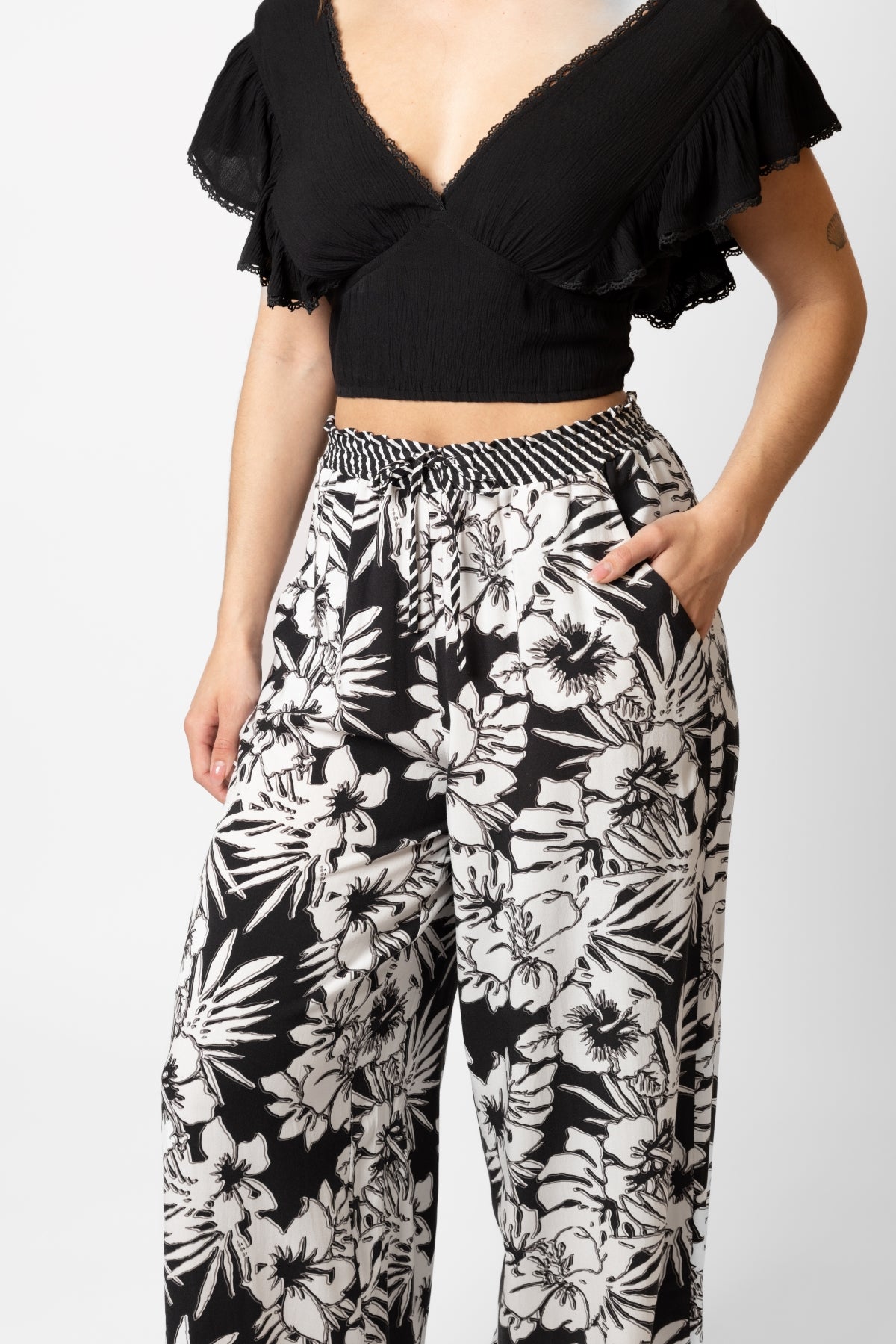 Brunette model facing front close up, wearing Black Onyx Copacabana Floral Wide Leg Pant. Features a vibrant hibiscus floral print, wide leg palazzo style, and an adjustable cinched waist with pockets. Crafted from 100% sand-washed rayon, perfect for a garden-chic look or vacation style. Koy Resort affordable vacation, cruise, and resort-wear.