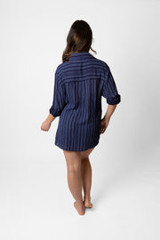 Brunette model facing back, wearing a navy Marina Stripe Relaxed Button Up Shirt. Features a relaxed fit, collar, white tone buttons, roll-up sleeves, and a tonal mock stitch print. Perfect for any occasion, combining style and comfort. Koy Resort affordable vacation, cruise, and resort-wear.