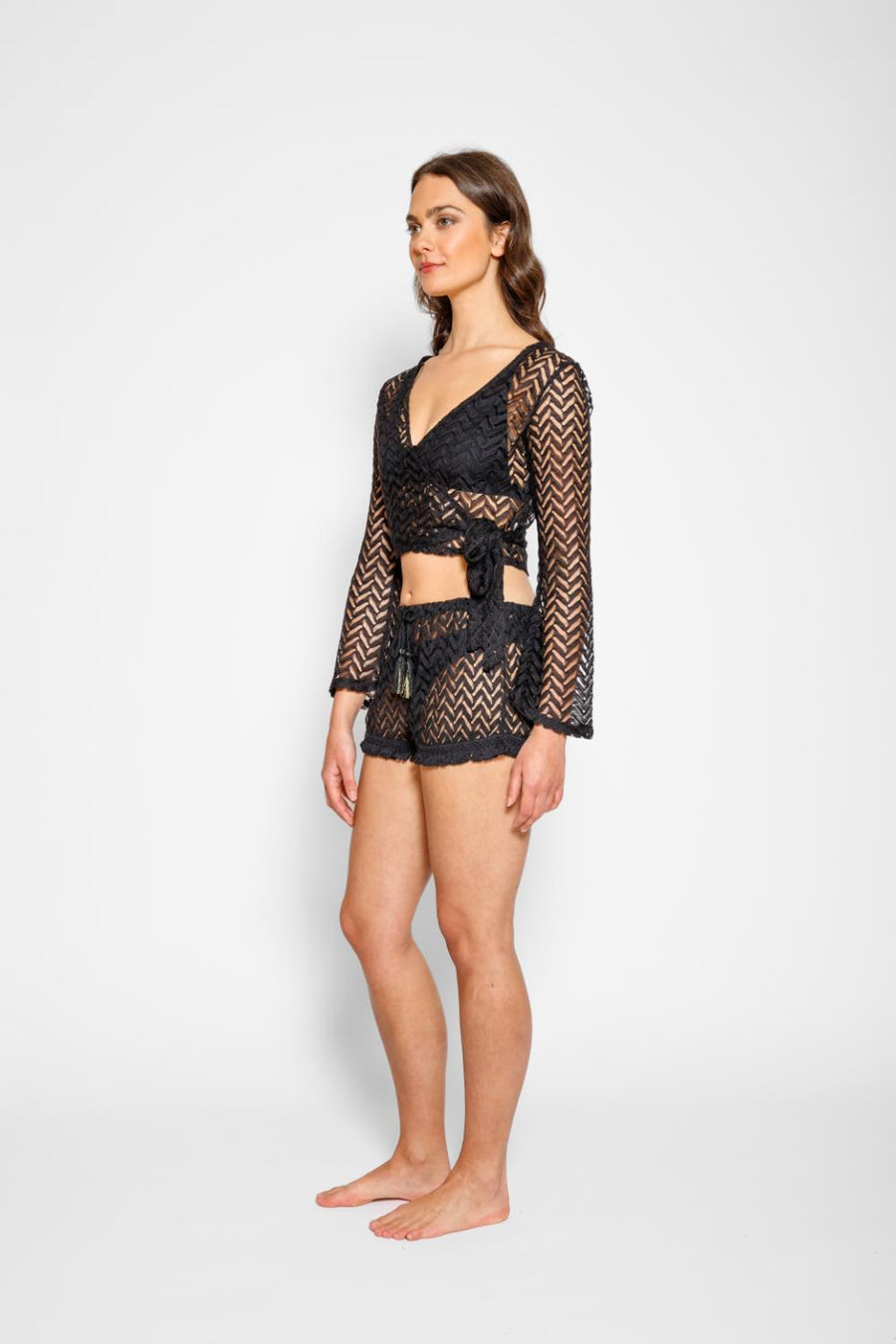 Brunette model facing side, wearing Black Zuma Long Sleeve Crochet Wrap Top with sheer crochet herringbone pattern and long bell sleeves. Versatile wrap design available in natural white or black. Perfect for a sassy cover-up look. Koy Resort affordable vacation, cruise, and resort-wear.