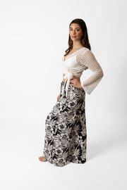 Brunette model facing side, wearing Black Onyx Copacabana Floral Wide Leg Pant. Features a vibrant hibiscus floral print, wide leg palazzo style, and an adjustable cinched waist with pockets. Crafted from 100% sand-washed rayon, perfect for a garden-chic look or vacation style. Koy Resort affordable vacation, cruise, and resort-wear.