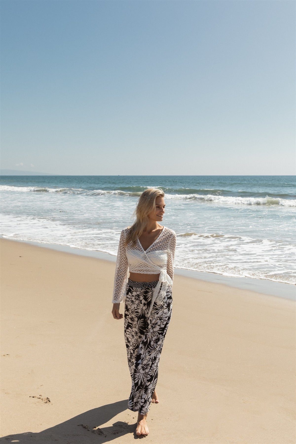 Blonde model facing front, wearing White Zuma Long Sleeve Crochet Wrap Top with sheer crochet herringbone pattern and long bell sleeves. Versatile wrap design available in natural white or black. Perfect for a sassy cover-up look. Koy Resort affordable vacation, cruise, and resort-wear.