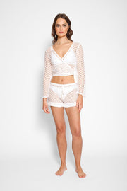 Brunette model facing front, wearing White Zuma Long Sleeve Crochet Wrap Top with sheer crochet herringbone pattern and long bell sleeves. Versatile wrap design available in natural white or black. Perfect for a sassy cover-up look. Koy Resort affordable vacation, cruise, and resort-wear.