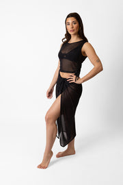 Brunette model facing side, wearing Black Seascape Mesh Long Sarong with a dark tonal floral print, adjustable size, midi length, and sheer fabric. Koy Resort affordable vacation, cruise, and resort-wear.