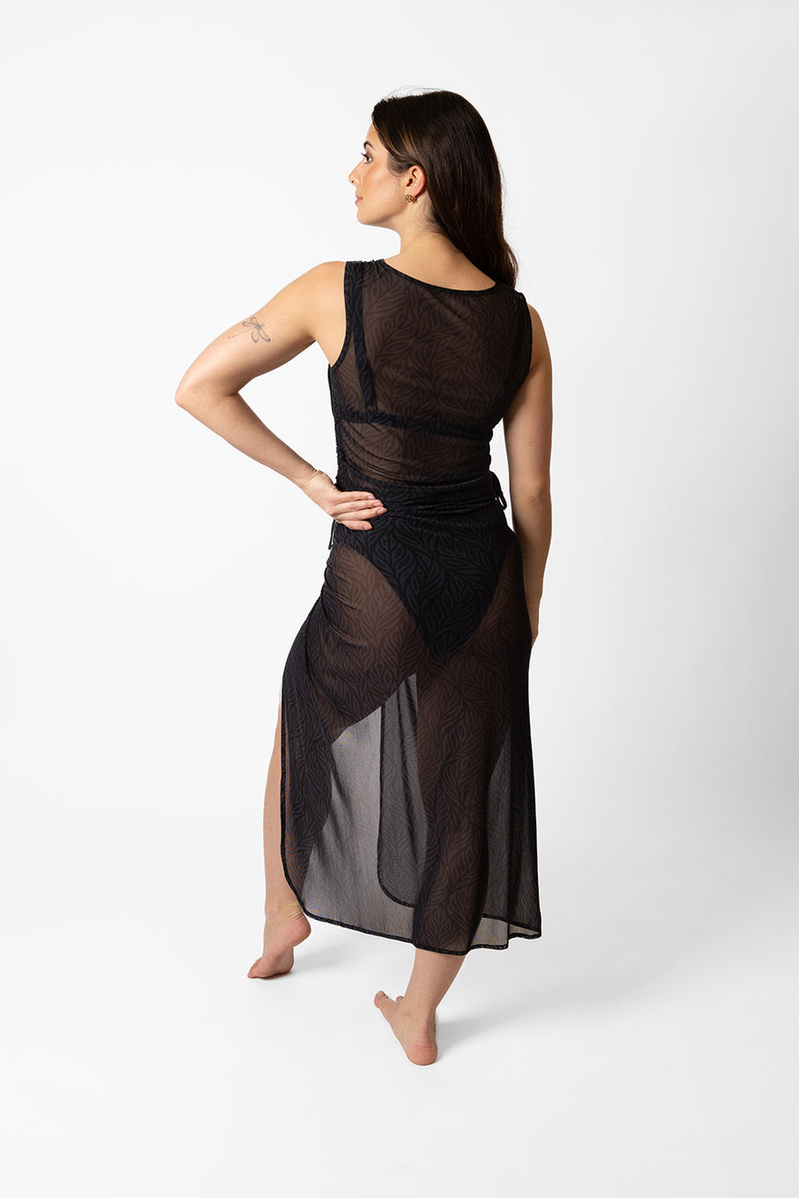 Brunette model facing back, wearing Black Seascape Mesh Long Sarong with a dark tonal floral print, adjustable size, midi length, and sheer fabric. Koy Resort affordable vacation, cruise, and resort-wear.