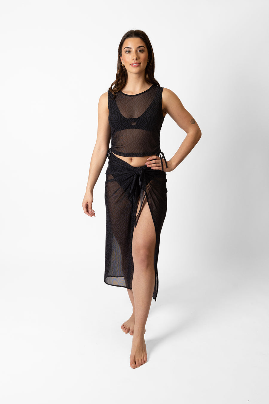 Brunette model facing front, wearing Black Seascape Mesh Long Sarong with a dark tonal floral print, adjustable size, midi length, and sheer fabric. Koy Resort affordable vacation, cruise, and resort-wear.