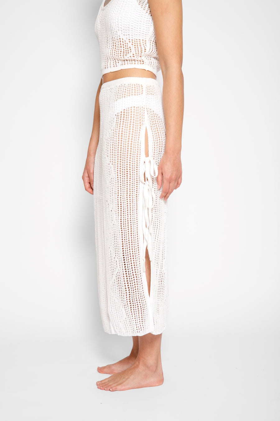 Brunette model facing side close up, wearing Cream Santorini Knit Midi Skirt with high side slits, adjustable ties, and semi-sheer fabric. Koy Resort affordable vacation, cruise, and resort-wear.