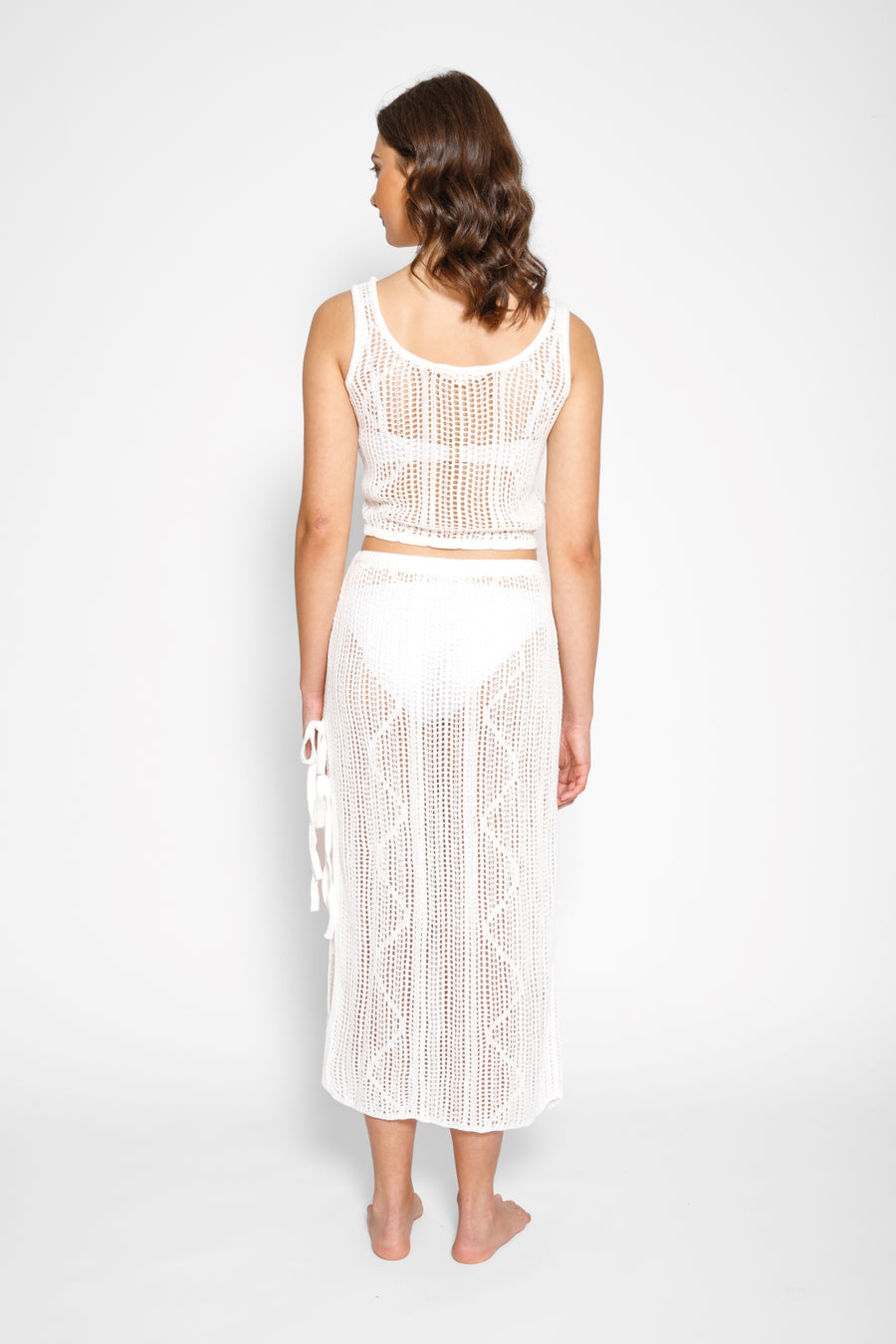 Brunette model facing back, wearing Cream Santorini Knit Midi Skirt with high side slits, adjustable ties, and semi-sheer fabric. Koy Resort affordable vacation, cruise, and resort-wear.