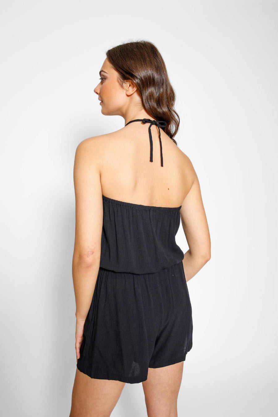 Brunette model facing back, wearing Black Miami Romper with an elastic top, adjustable waist with a tassel detail, elastic waist, and pockets. Koy Resort affordable vacation, cruise, and resort-wear