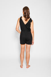 Brunette model facing back, wearing Black Laguna Beach Romper, made of soft jersey spandex fabric with adjustable straps and pockets. Koy Resort affordable vacation, cruise, and resort-wear