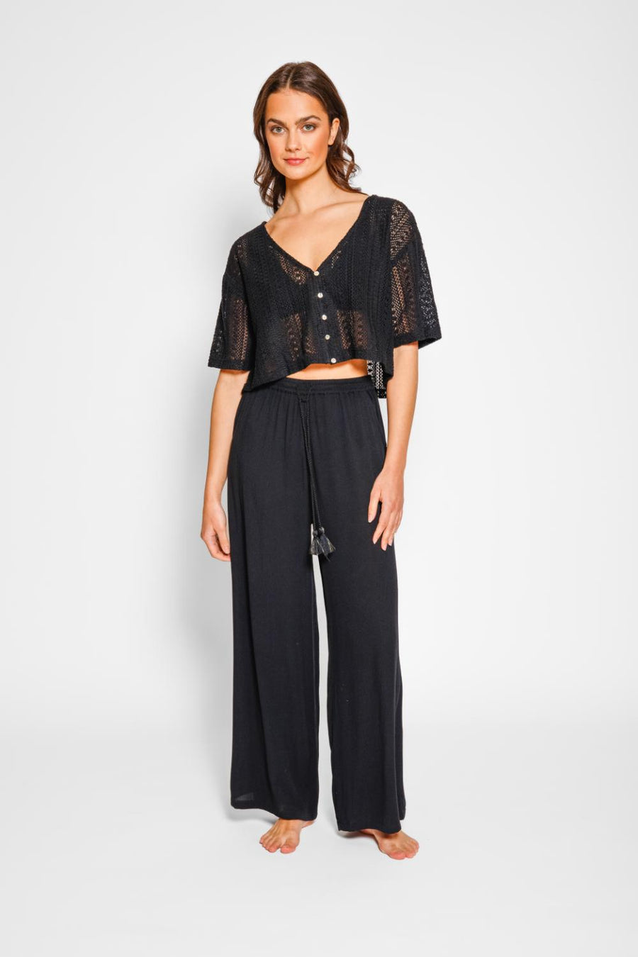 Brunette model facing front, wearing Black Miami Wide Leg Pant with a tie waist, palazzo style, flowy straight leg, elastic waist, side pockets, and metallic tassel drawcord. Koy Resort affordable vacation, cruise, and resort-wear.