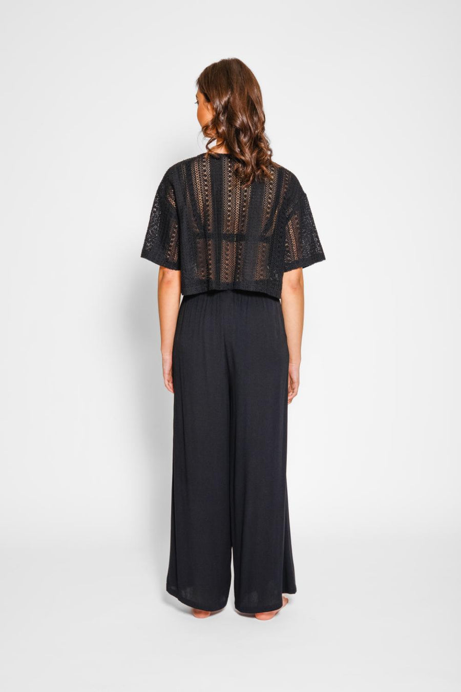 Brunette model facing black, wearing Black Miami Wide Leg Pant with a tie waist, palazzo style, flowy straight leg, elastic waist, side pockets, and metallic tassel drawcord. Koy Resort affordable vacation, cruise, and resort-wear.