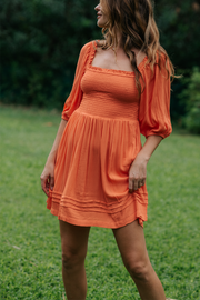 Blonde model facing side, wearing Orange Coral Punch Miami Smocked Mini Dress. Features a smocked bodice for a figure-flattering fit and puffy peasant-style sleeves for a bohemian touch. Perfect for the beach, a summer wedding, or a night out. Koy Resort affordable vacation, cruise, and resort-wear.