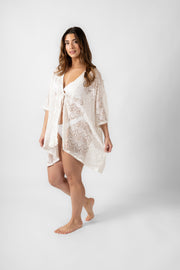 Brunette model facing front, wearing the White Flamenco Lace Crochet Beach Cover Up Kimono. This stylish cover-up features an open front, scoop hem, long tie-up tassel, short sleeves, and sheer crochet lace detailing. Made from 55% Cotton and 45% Nylon. Koy Resort affordable vacation, cruise, and resort-wear.