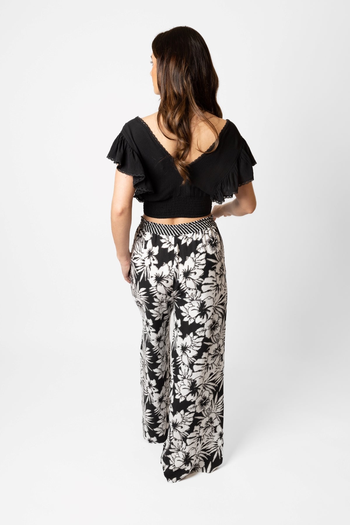 Brunette model facing back, wearing Black Onyx Copacabana Floral Wide Leg Pant. Features a vibrant hibiscus floral print, wide leg palazzo style, and an adjustable cinched waist with pockets. Crafted from 100% sand-washed rayon, perfect for a garden-chic look or vacation style. Koy Resort affordable vacation, cruise, and resort-wear.