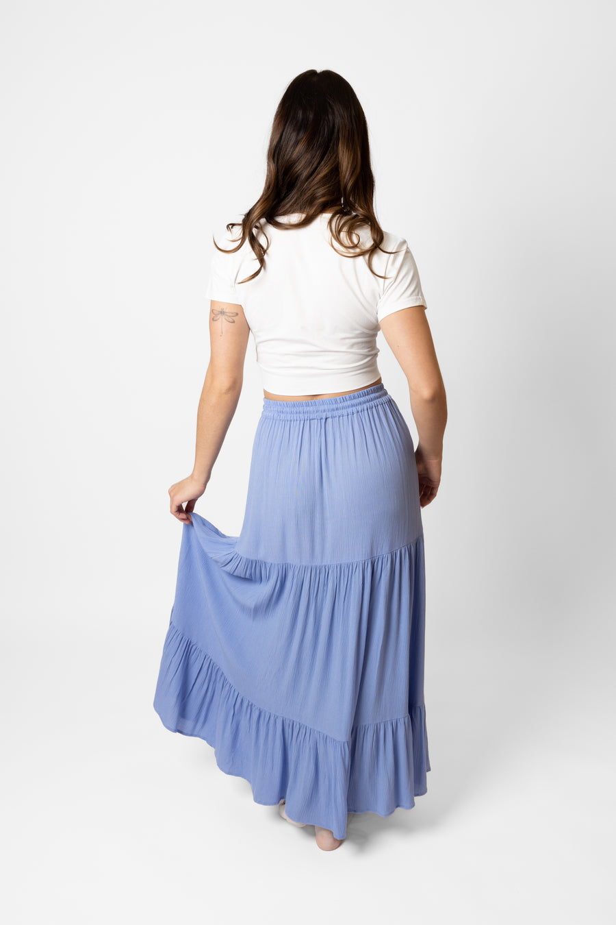 Brunette model facing back, wearing Bahama Blue Miami Tiered Boho Maxi Skirt with a flowy fit, adjustable elastic waistband and beaded drawcord, pockets, and white lining. Made from crinkle rayon. Perfect for bohemian beach days and romantic getaways. Koy Resort affordable vacation, cruise, and resort-wear.