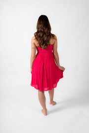 Brunette model facing back, wearing Raspberry Pink Miami Strapless Bandeau Mini Dress. This loose and flowy dress features an elastic bandeau top with a peek-a-boo bodice, and side slits for a flirty look. The dress is lined in white and has an adjustable cord at the back. Koy Resort affordable vacation, cruise, and resort-wear.