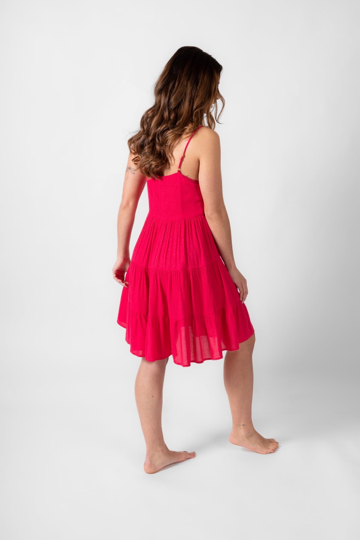 Brunette model facing back, wearing Raspberry Pink Miami Tiered Strappy Mini Dress. The dress features adjustable straps, a flirty tiered empire silhouette, and is crafted from lightweight rayon for a comfortable feel. It includes a smocked back, is lined in white, and has pockets, making it both stylish and practical. Koy Resort affordable vacation, cruise, and resort-wear.