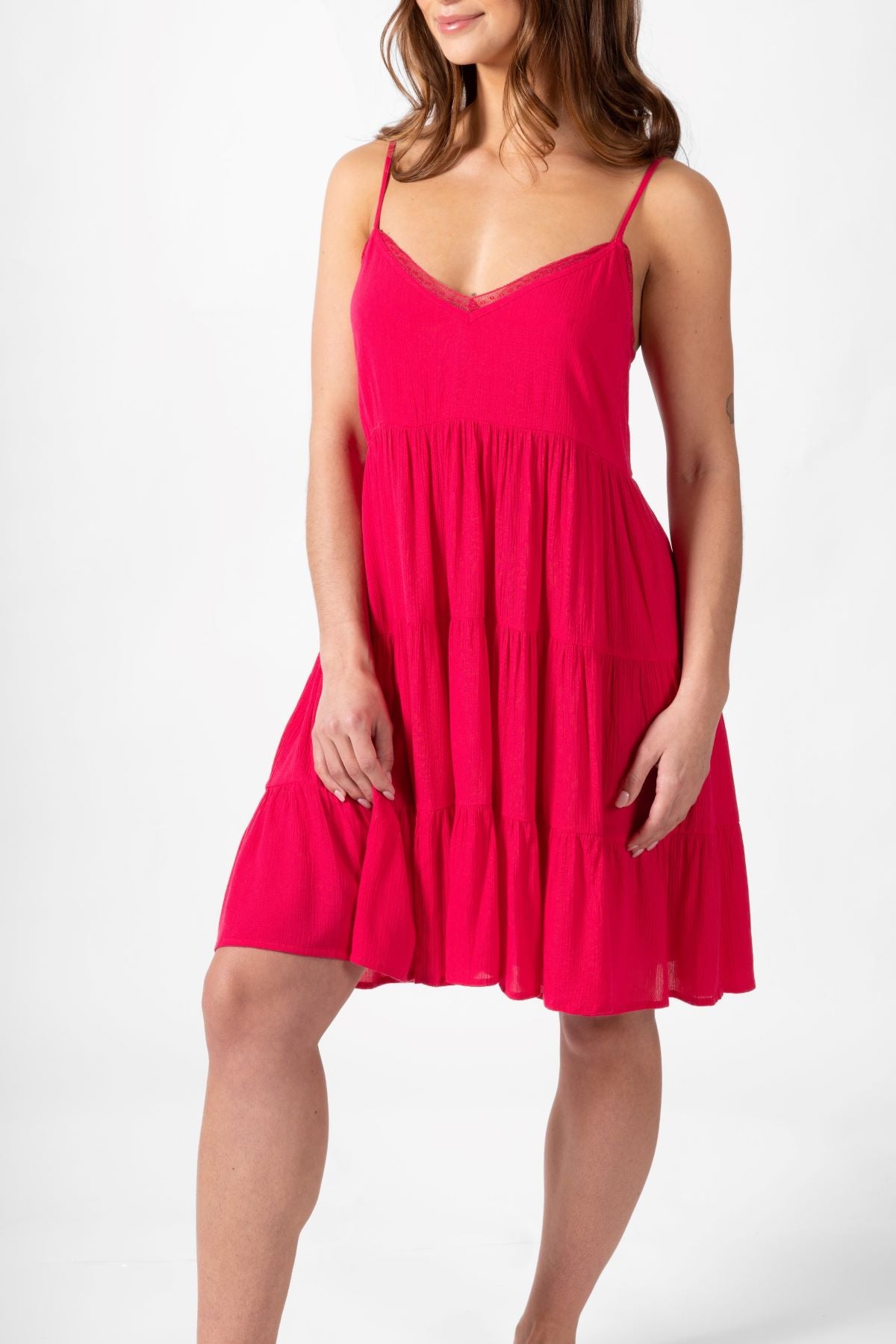 A front shot of a brunette hair woman holding the edge of her raspberry colored spaghetti strap summer beach dress. Brunette model facing front side close up, wearing Raspberry Pink Miami Tiered Strappy Mini Dress. The dress features adjustable straps, a flirty tiered empire silhouette, and is crafted from lightweight rayon for a comfortable feel. It includes a smocked back, is lined in white, and has pockets, making it both stylish and practical. Koy Resort affordable vacation, cruise, and resort-wear.