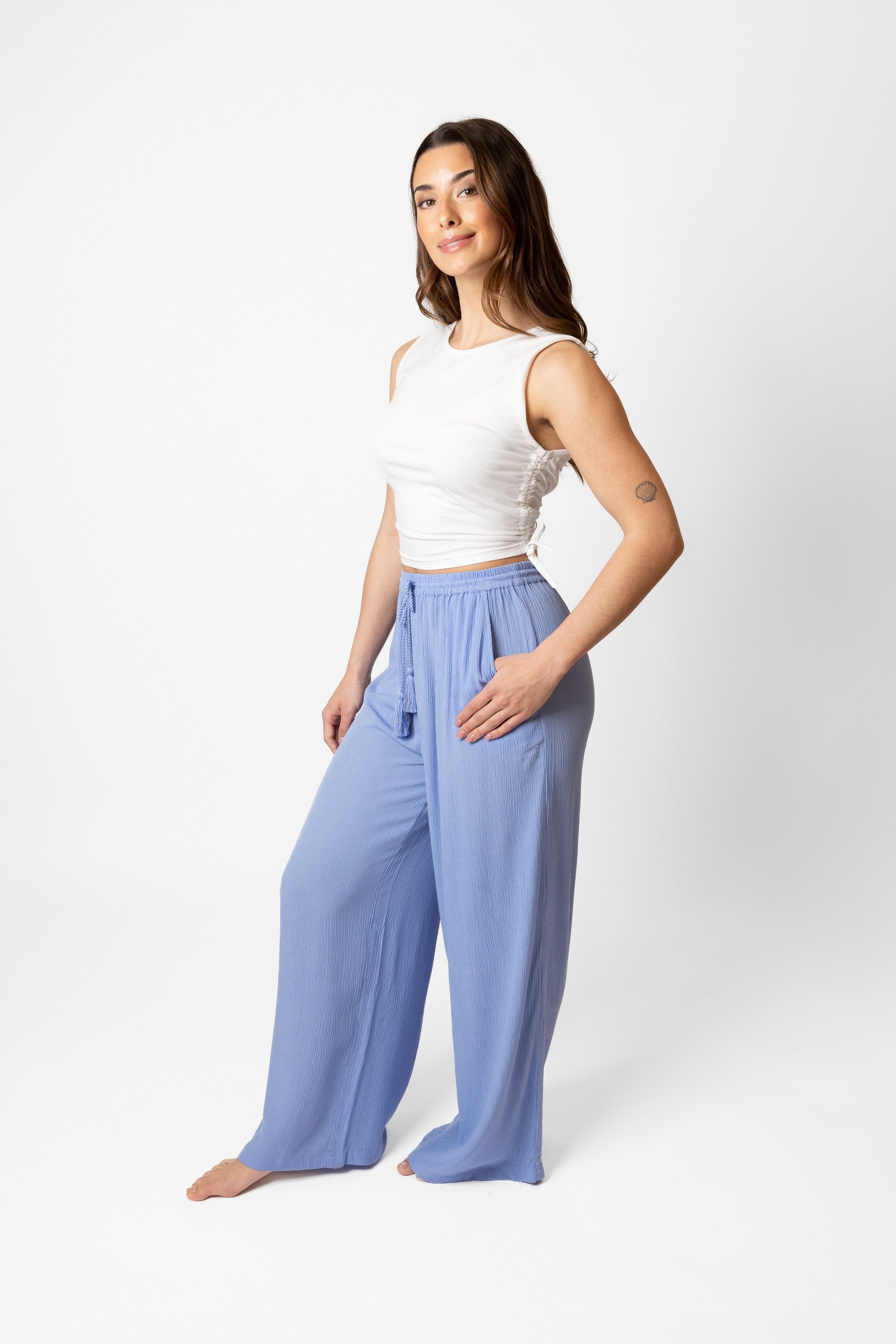 Brunette model facing side, wearing Bahama Blue Miami Wide Leg Pant with a flowy straight leg, elastic waist, side pockets, metallic tassel drawcord, and white lining. Palazzo style for a fun and flattering fit. Koy Resort affordable vacation, cruise, and resort-wear.