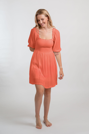 Blonde model facing front, wearing Orange Coral Punch Miami Smocked Mini Dress. Features a smocked bodice for a figure-flattering fit and puffy peasant-style sleeves for a bohemian touch. Perfect for the beach, a summer wedding, or a night out. Koy Resort affordable vacation, cruise, and resort-wear.sort-wear.