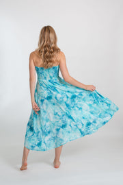 A woman wearing Koy Resort Aquarelle collection blue tie-dye dress one hand holding the edge of the dress, lifting it slightly. The dress is a vibrant shade of blue, with a unique tie-dye pattern that creates a marbled effect. The dress is sleeveless and appears to have a flowy, relaxed fit. 