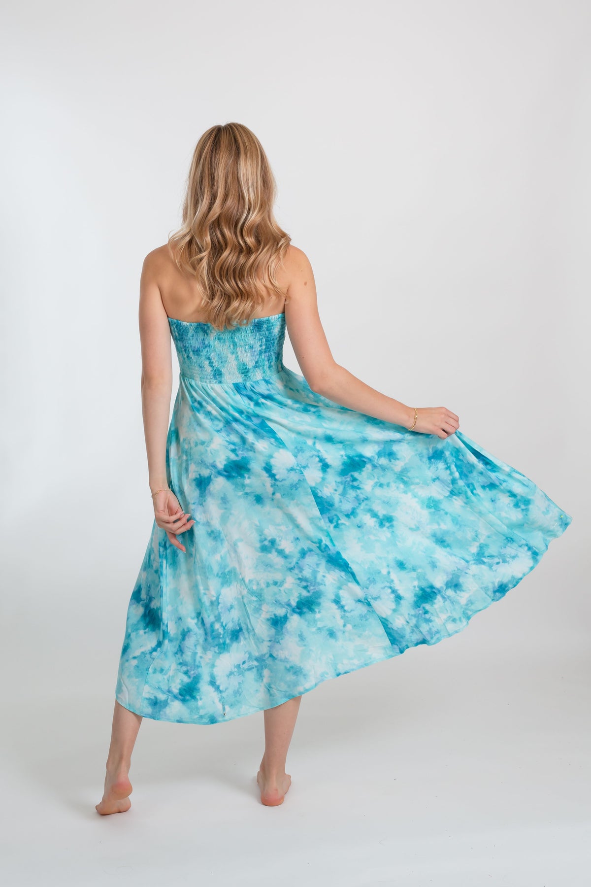 A woman wearing Koy Resort Aquarelle collection blue tie-dye dress one hand holding the edge of the dress, lifting it slightly. The dress is a vibrant shade of blue, with a unique tie-dye pattern that creates a marbled effect. The dress is sleeveless and appears to have a flowy, relaxed fit. 