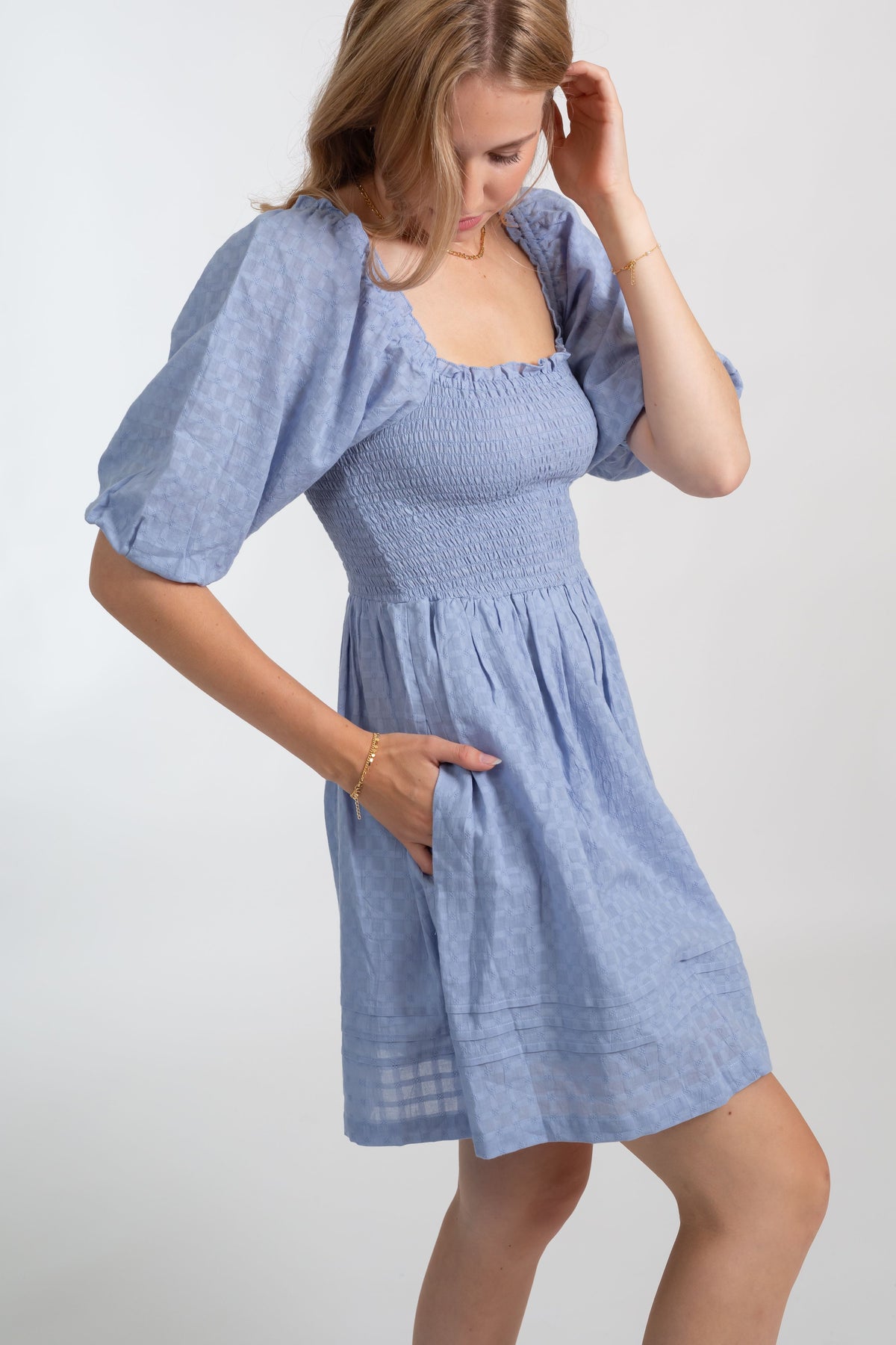 a zoomed in photo of a blonde hair female model wearing a sky blue color short sleeve light weight mini dress with one hand in her pocket of the dress
